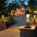 Antique gold solar post cap light glowing warm white on a stone pillar at dusk outside a large home