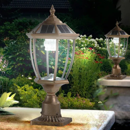 Antique gold solar pillar light lit in a garden setting with flowers and greenery behind