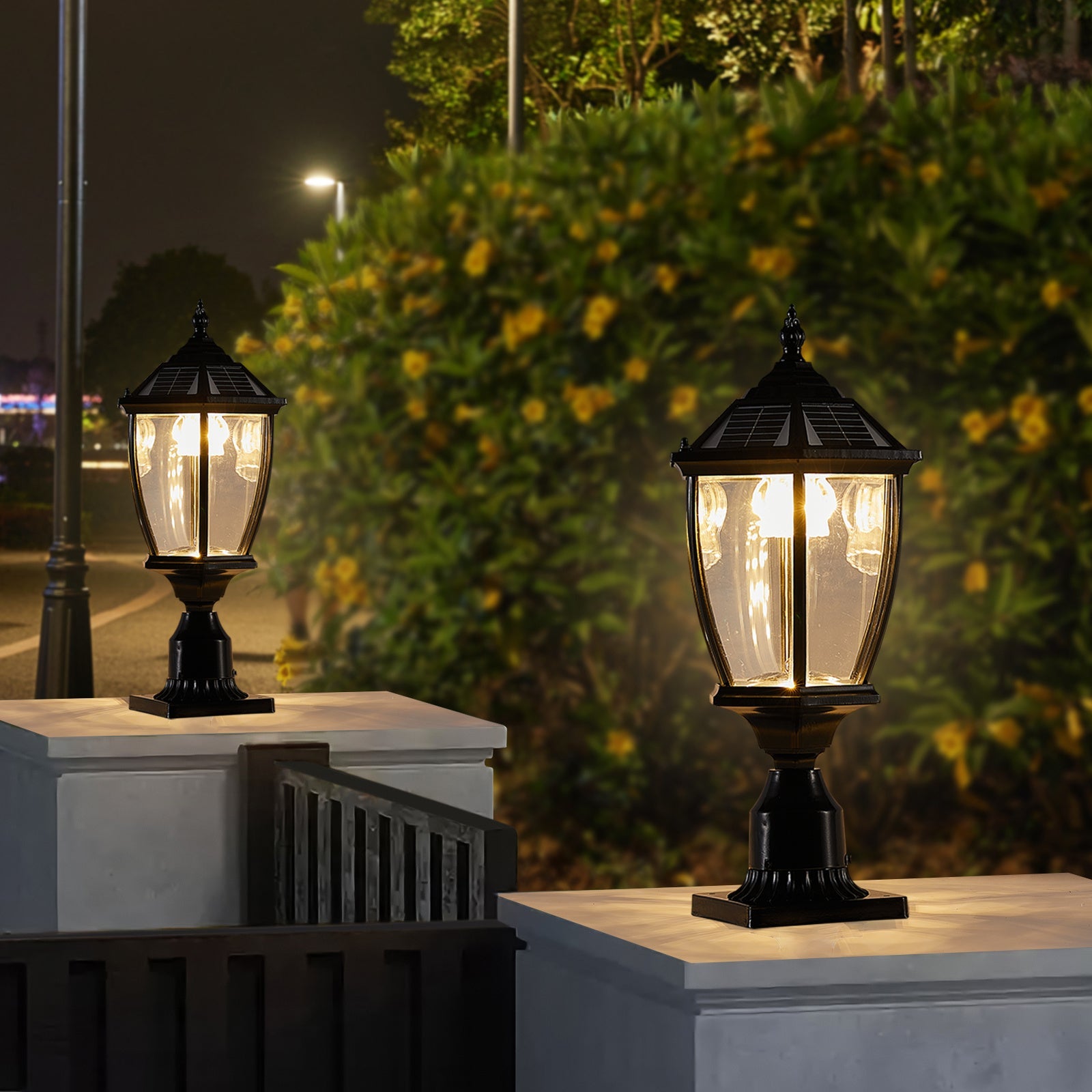 Two ornate Victorian solar pillar lights glowing warm white at night beside a flowering hedge