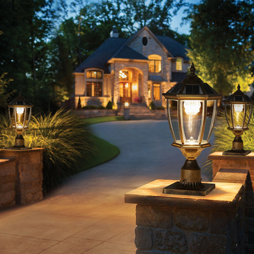 Antique gold solar post cap lights glowing warm white on stone pillars at dusk, lining a driveway in front of a large stone house