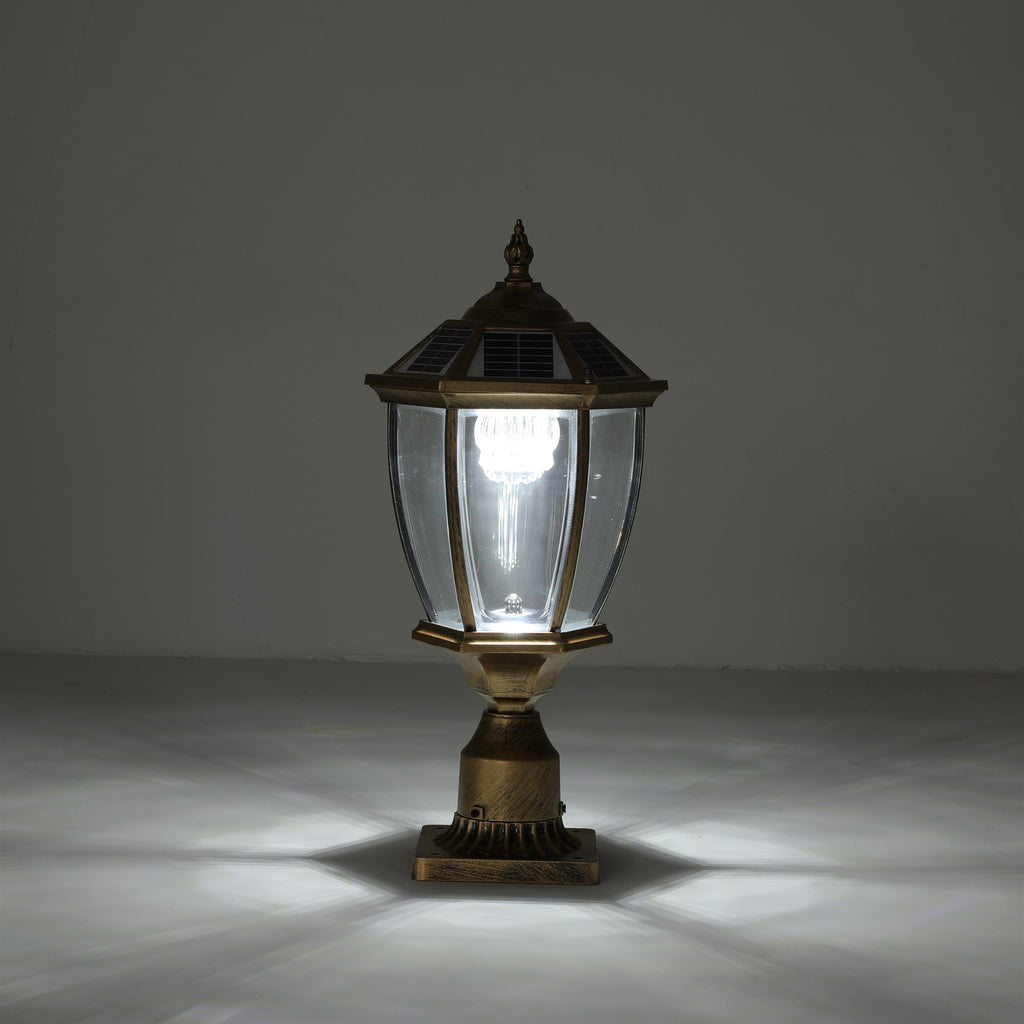 Antique gold solar post cap light lit with a warm glow on its pedestal base, showing the full hexagonal lantern, solar cap, fluted metal base, and light pattern cast on the floor