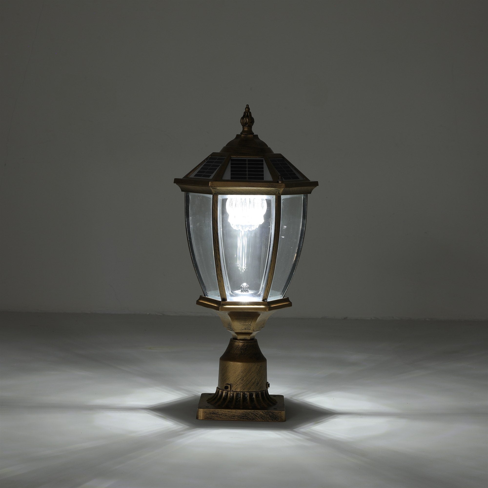 Antique gold solar post cap light lit with a warm glow on its pedestal base, showing the full hexagonal lantern, solar cap, fluted metal base, and light pattern cast on the floor
